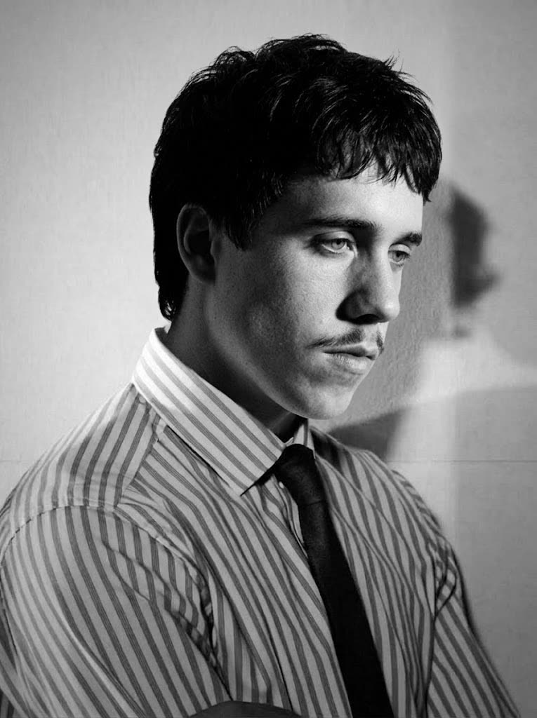 Ollie Bennett, photographed in black and white against a plain pale wall,
                turned in three-quarter view with his gaze lowered and away from the camera.